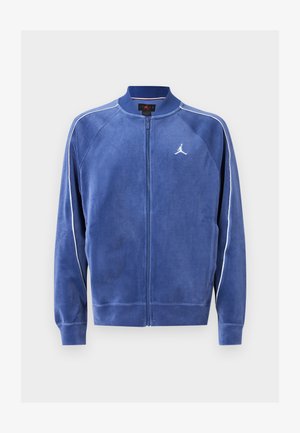 Casaco azul com fecho éclair, feito de material suave. Apresenta acabamentos brancos nas mangas e um logótipo no peito. Gola e punhos canelados.