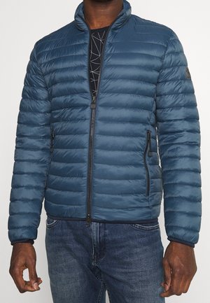 Mężczyzna w niebieskiej pikowanej kurtce zapinanej na zamek, czarnej koszuli w wzory i niebieskich dżinsach, stojący na jednolitym tle.