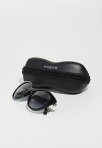 Ochelari de soare negri cu rame supradimensionate în stil cat-eye, lentile gri și accente aurii, alături de un etui negru cu fermoar cu logo-ul "vogue".