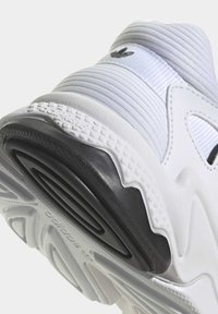 Vitt sportskor med texturerad ovandel, med svart yttersula och zigzagmönstrad sula. Tydlig logodetalj vid hälen.