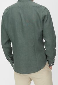 Zielona lniana koszula z długim rękawem, kołnierzykiem na guziki, widocznymi szwami i zapinanymi na guziki mankietami. Tył ma zakrzywiony dół oraz widoczną fakturę tkaniny.