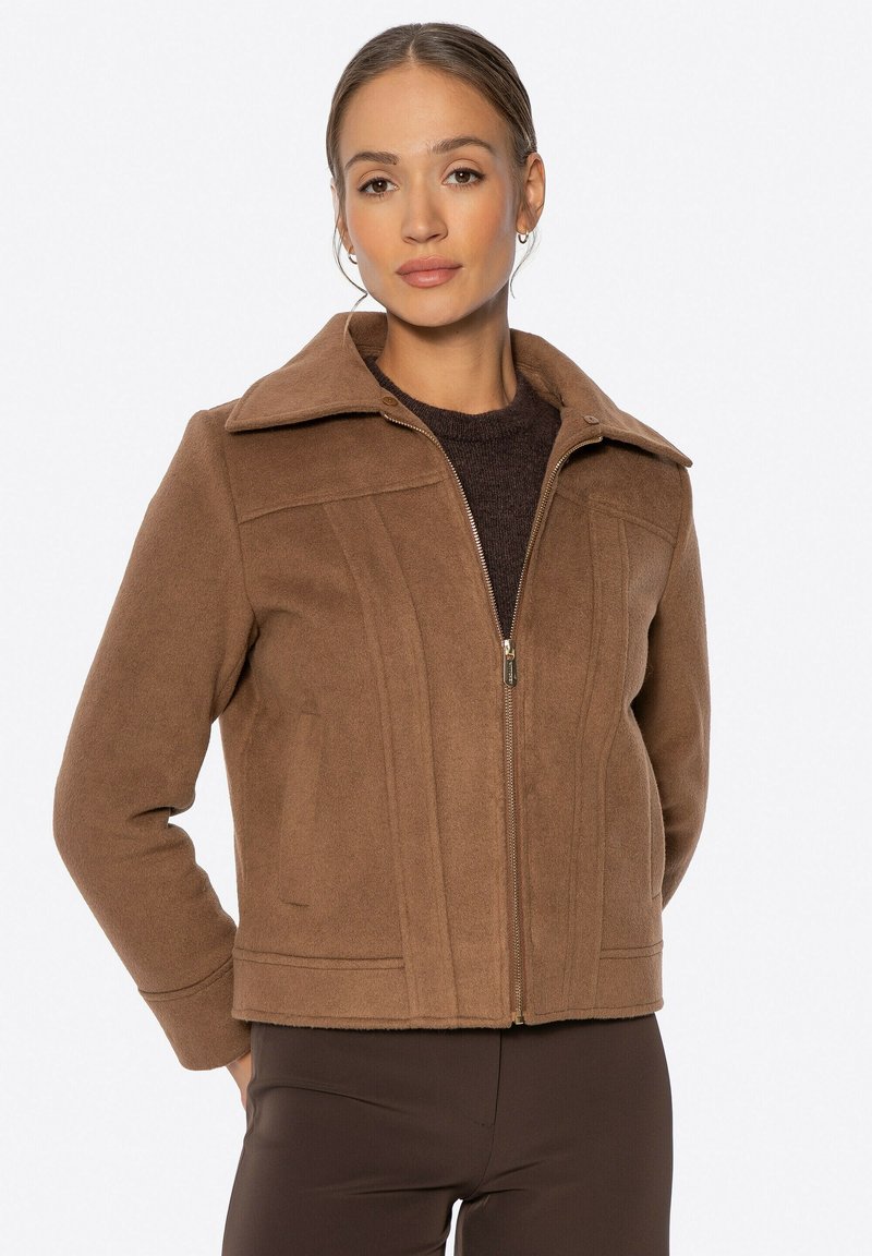 Giacca in misto lana marrone con colletto, zip frontale, cuciture strutturate e una vestibilità leggermente cropped. Il tessuto ha una consistenza morbida.