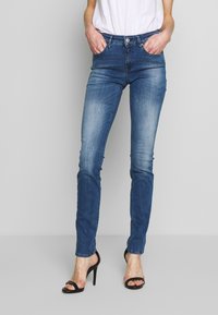 Jeans ajustados de color azul, confeccionados en denim, con un acabado desgastado, cinco bolsillos y cierre de botón. Combinados con sandalias de tacón alto negras.