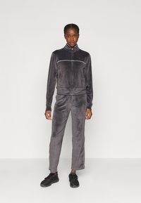 Tuta in velluto grigio con giacca con zip e pantaloni con vita elastica, caratterizzata da strisce laterali bianche e un piccolo logo sul petto.