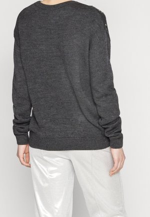 Person iført en mørkegrå strikket sweater med knapdetaljer på skulderen og lyse, skinnende bukser, vist bagfra.