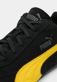 Sneaker in suede nero con accenti gialli; presenta lacci neri piatti, superficie testurizzata e marchio Puma sul lato e sulla linguetta.