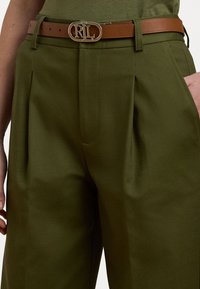 Calças ajustadas verde-oliva com pregas, apresentando um cinto de pele castanho com uma fivela prateada com o logótipo 'RL'. Tecido liso, sem padrões.