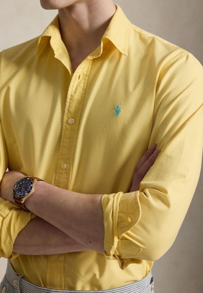 Personne portant une chemise jaune à manches longues boutonnée avec un logo bleu brodé et une montre au poignet gauche, bras croisés.