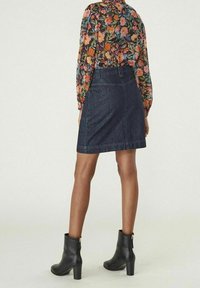 Kvinna klädd i en mörk jeanskjol, blommig blus med långa ärmar och svarta högklackade ankelboots stående mot en enfärgad bakgrund.