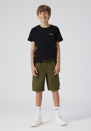 Smilende dreng iført sort T-shirt, olivengrønne cargoshorts, hvide sokker og hvide sneakers, stående mod en ensfarvet lys baggrund.