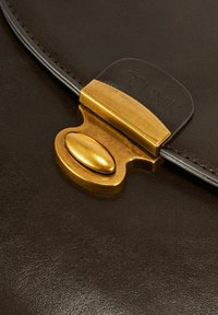 Zbliżenie na ciemnobrązową skórzaną torbę z błyszczącą złotą klamrą i teksturowaną powierzchnią, z czarnym paskiem przymocowanym do torby.