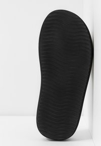 Semelle de chaussure noire avec motif de bande de roulement ondulé texturé, positionnée verticalement contre un mur d’angle blanc.