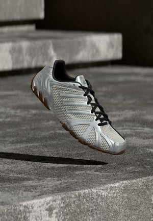 Srebrno-crna Adidas sportska cipela s teksturiranim dizajnom i smeđim đonom, visi iznad sive betonske površine i stepenica.