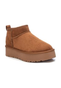 Botas de tobillo de ante marrón con punta redonda, un cuello bajo y una suela gruesa color beige. Presentan costuras mínimas y una pestaña en la parte posterior.