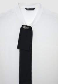 Blusa bianca con dettaglio di fiocco nero impreziosito da un cristallo sul collo. Tessuto liscio con chiusura a bottoni, design elegante.