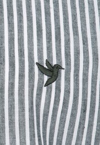 Una tela a rayas en gris y blanco presenta un pequeño colibrí bordado en verde. La textura parece tejida y el patrón es consistente.