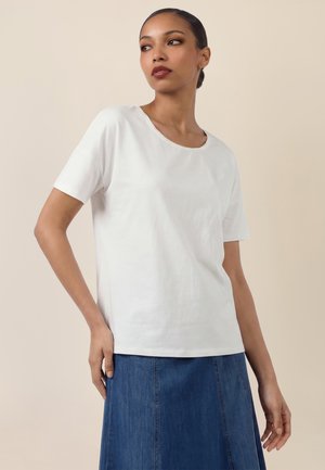 Woman wearing a plain white short-sleeve shirt and a blue denim skirt, standing with one hand on her hip against a beige background.