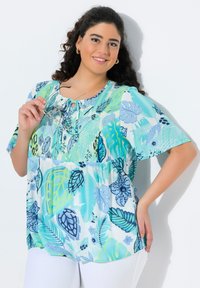 Blusa leggera a manica corta con un motivo di foglie blu e verdi, dettagli con bottoni e scollatura rotonda. Indossata con pantaloni bianchi.