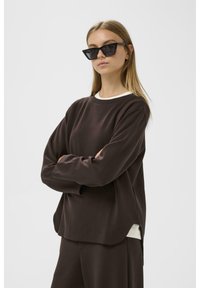 Sudadera marrón oscuro con cuello redondo y mangas largas, que presenta un ribete blanco en el escote y un ajuste holgado. Llevada con gafas de sol de ojo de gato negras.