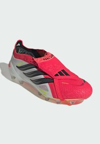 Ποδοσφαιρικό παπούτσι Adidas Predator σε κόκκινο και γκρι χρώμα με μαύρες ρίγες, σύστημα κορδονιών που καλύπτονται και διάφανα πέλματα σε λευκό φόντο.