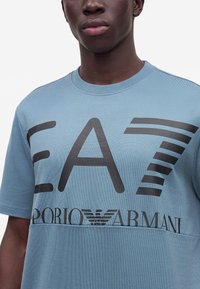 Νεαρός άνδρας που φοράει ένα ανοιχτό μπλε t-shirt Emporio Armani EA7 με έντονα μαύρα γράμματα και λογότυπο στο στήθος.