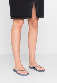 Grå flip-flops med en tekstureret vævet sål og glatte remme, placeret på bare fødder, parret med en sort denimnederdel.