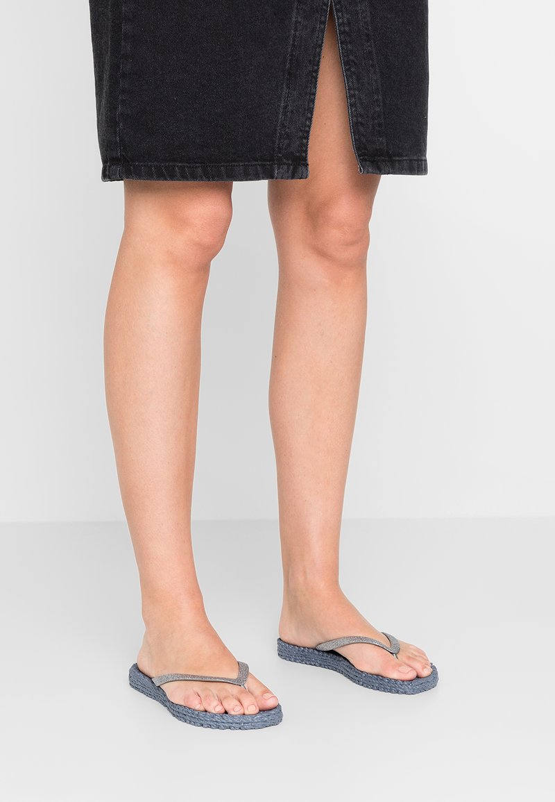 Grå flip-flops med en tekstureret vævet sål og glatte remme, placeret på bare fødder, parret med en sort denimnederdel.