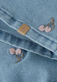 Sininen farkkukangas, jossa on kirjailtu vaaleanpunaisia kirsikoita ja saumassa ommeltu pieni ruskea nahkainen merkki.