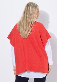 Röd stickad poncho med korta ärmar, ribbad struktur, öppna sidor och en avslappnad passform, lagd över en vit skjorta.