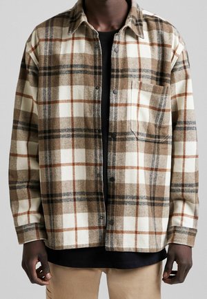 Mann trägt ein braun-beige-cremefarbenes kariertes Hemd über einem schwarzen T-Shirt und beigen Hosen, steht mit entspannten Armen.