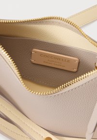 Lys beige skinnveskeinnside med glidelås i gull og kremfarget fôr. Coccinelle-merke preget på lysbrun skinnmerkelapp. Strukturert overflate.