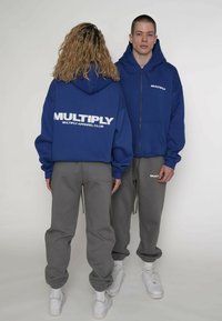 Felpa con cappuccio blu con cerniera e scritta bianca "MULTIPLY" sul retro, abbinata a pantaloni della tuta grigi e sneaker bianche, indossata da due modelli visti da dietro.
