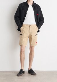 Pantalones cortos cargo beige con bolsillos con cremallera, combinados con una camisa negra de botones y una camiseta blanca, completados con zapatillas negras.