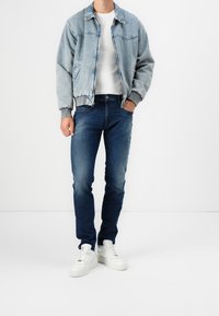 Chaqueta de mezclilla azul clara con puños grises, combinada con jeans azul oscuro y zapatillas blancas. La chaqueta cuenta con cierre de cremallera y bolsillos.