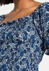 Blå blommig topp med smockad byst och korta ärmar. Har en blandning av mörkblå och ljusblå blommor på en vit bakgrund.