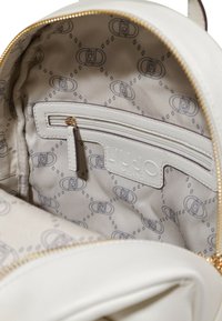 Interno di uno zaino in pelle bianca con una tasca con zip, logo impresso e fodera decorata con un motivo ripetuto in grigio.