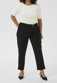 Femme portant un haut blanc ajusté à manches courtes, un pantalon noir cropped taille haute avec ceinture, et des escarpins noirs à talon ouvert, debout.