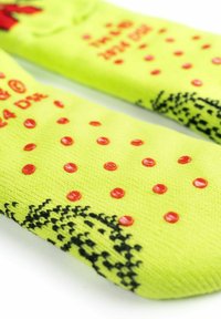 Calzini giallo brillante con puntini rossi antiscivolo in rilievo e talloni neri a fantasia, poggiati su una superficie bianca.