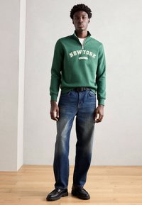 Sweatshirt vert avec l'impression "NEW YORK", col zippé ; porté avec un jean bleu droit et des mocassins noirs épais ; sol en bois et fond clair.