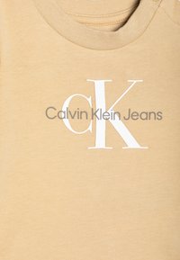 Hellbraunes Baumwoll-T-Shirt mit Rundhalsausschnitt, gut sichtbarem weißem "CK"-Logo und grauem "Calvin Klein Jeans"-Schriftzug darunter. Glatte Textur.