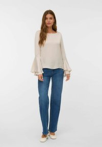 Blusa beige a maniche lunghe con polsini svasati, abbinata a jeans a gamba larga blu e scarpe di colore chiaro su uno sfondo neutro.