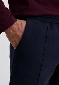 Marineblauwe broek met een gladde textuur, voorzien van een zijzak en een aansluitend ontwerp, aangevuld met een bordeauxrode langemouwtop in close-up weergave.