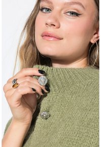 Junge Frau mit Flügel-Eyeliner, die silberne Knöpfe in Form eines Löwenkopfs an einem grünen Strickkleid trägt und den Kragen nahe an ihr Gesicht hält.