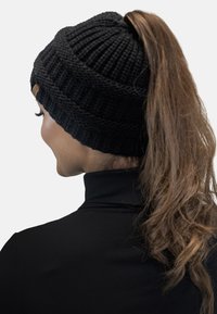 Zwarte gebreide beanie met een ribbelstructuur, voorzien van een zijlabel en geschikt voor een paardenstaart bovenaan. Eenvoudig, casual ontwerp.