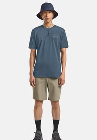 T-shirt da uomo a maniche corte color teal con un piccolo logo frontale, abbinato a pantaloni corti di colore khaki chiaro e un cappello a secchiello blu navy. Tessuto testurizzato e vestibilità casual.