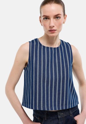 Femme à la peau claire et cheveux tirés en arrière, portant un haut bleu sans manches à rayures verticales blanches et un jean denim foncé, sur fond blanc.