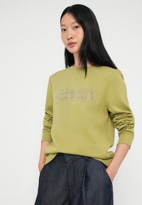 Πράσινη μπλούζα με μακριά μανίκια, στρογγυλή λαιμόκοψη, με μεταλλική γραφιστική λεπτομέρεια στο μπροστινό μέρος. Συνδυάζεται με σκούρα τζιν παντελόνια.