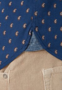 Chemise bleue avec petits motifs cachemire marron non rentrée sur un pantalon en velours beige, montrant les détails du tissu et un bouton.