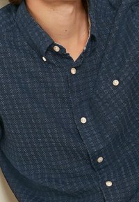 Camicia button-up blu navy con un motivo testurizzato, caratterizzata da accenti chiari, colletto e bottoni beige lungo la patta frontale.