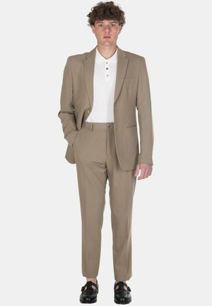 Giovane uomo con capelli ricci che indossa un completo beige, una camicia bianca con bottoni e scarpe eleganti nere, in piedi con una mano in tasca.